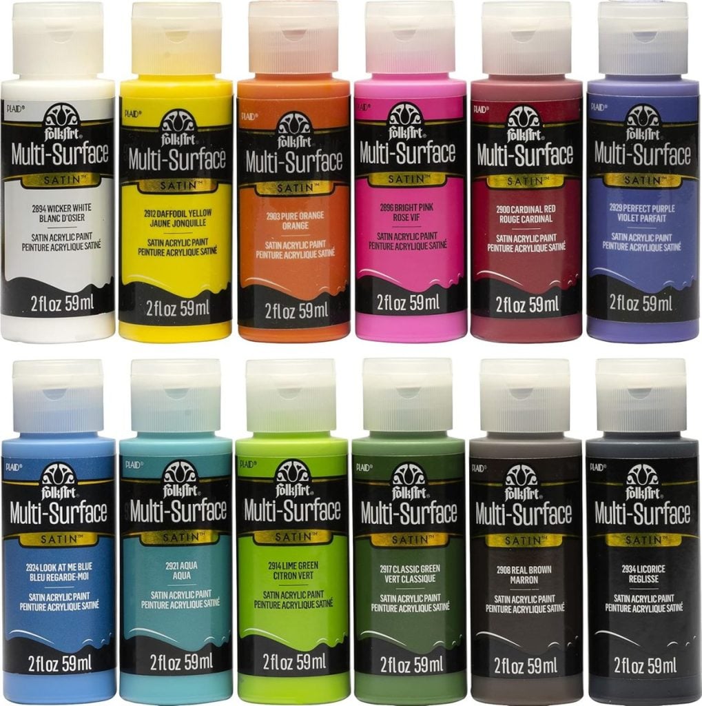 Twelve bottles of FolkArt Multi-Surface Satin acrylic paint in various colors are arranged in two rows on a white background.