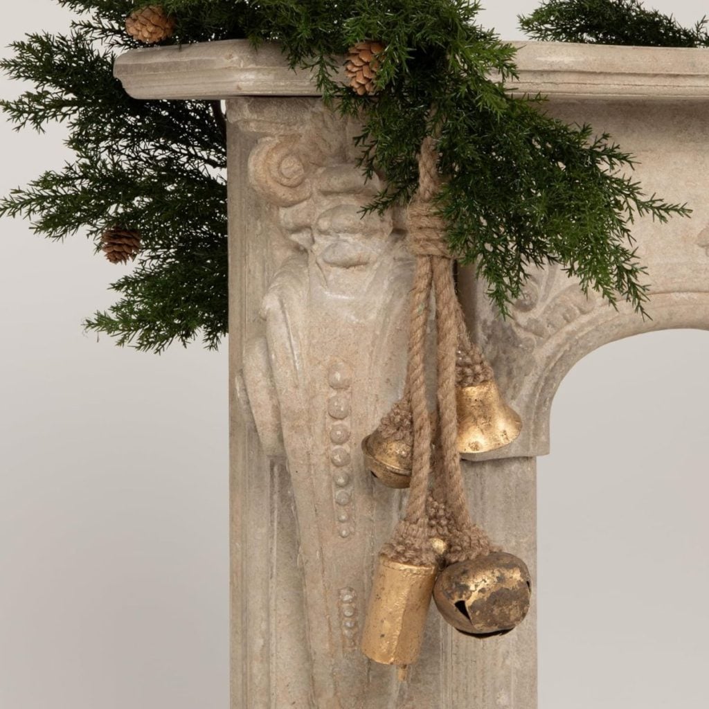 Stone mantel decorated with pine branches, small pinecones, and a rope with three gold bells hanging from the corner.