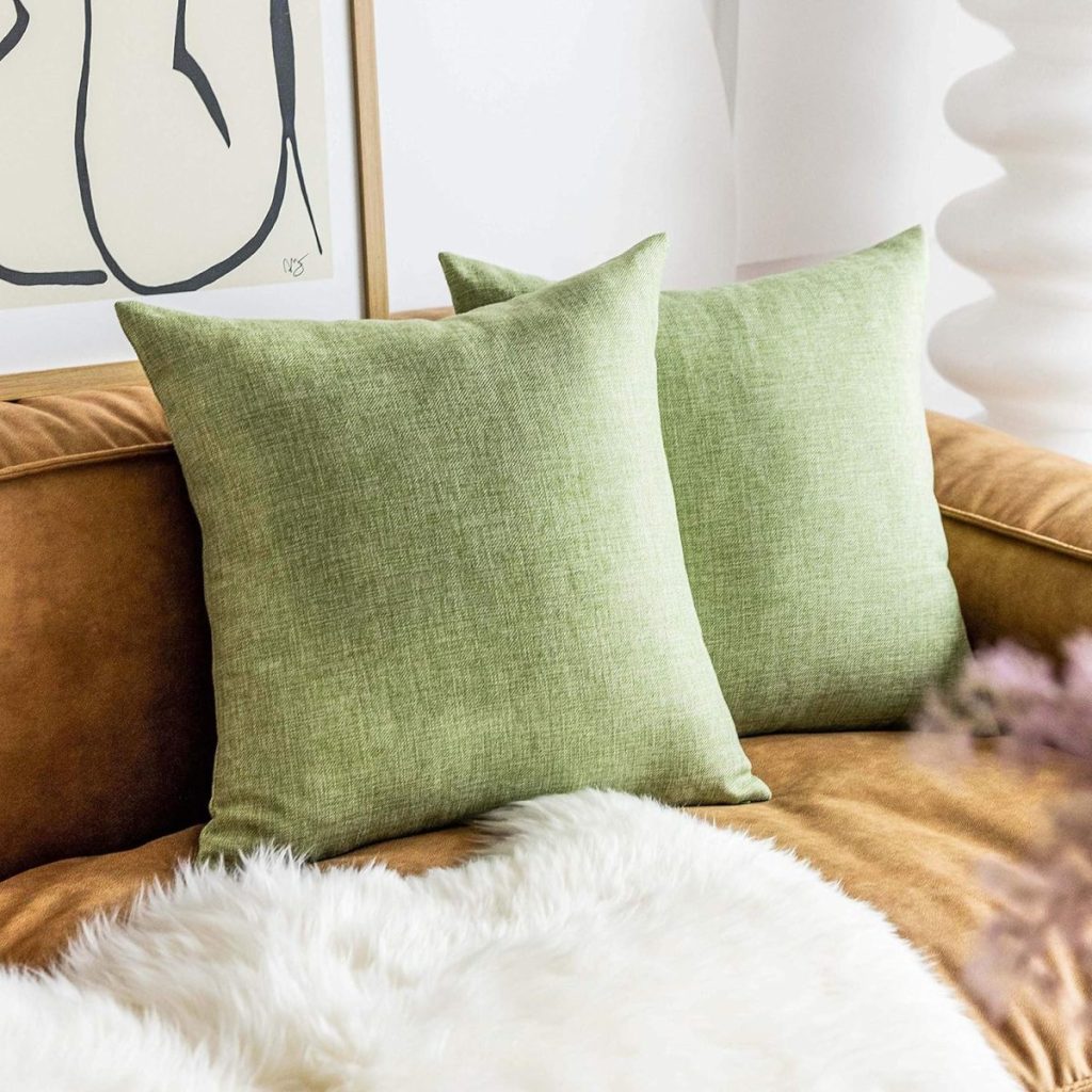 Two green throw pillows are placed on a brown sofa with a white faux fur blanket. A framed abstract artwork hangs in the background.