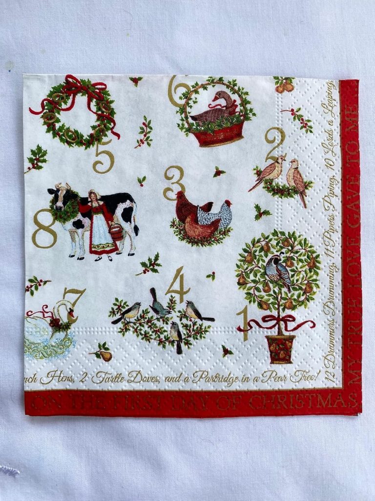 A decorative napkin featuring illustrations representing the 12 Days of Christmas song, with numbers, animals, birds, and festive wreaths in a holiday-themed design.