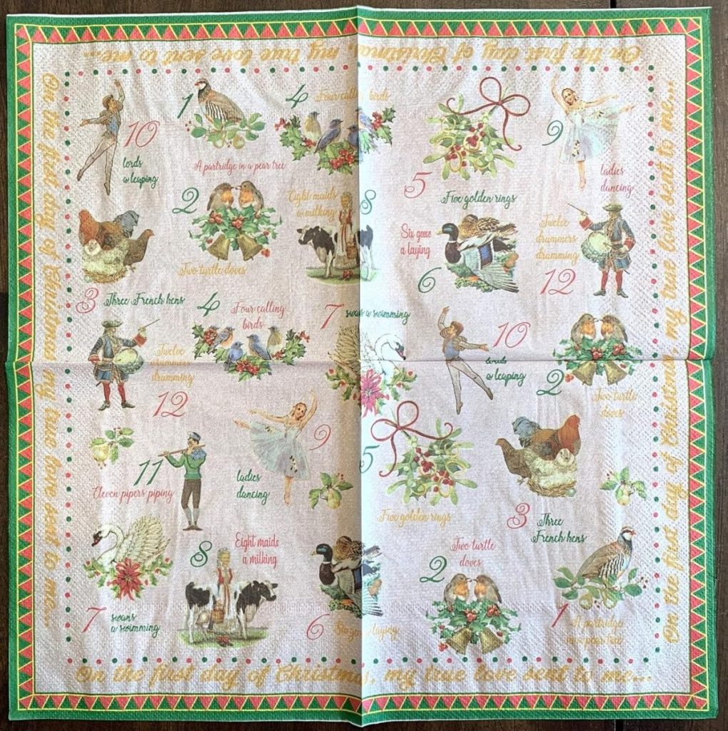 A decorative cloth featuring illustrations and text from the "Twelve Days of Christmas" song, showing numbers and corresponding festive images in a grid layout.
