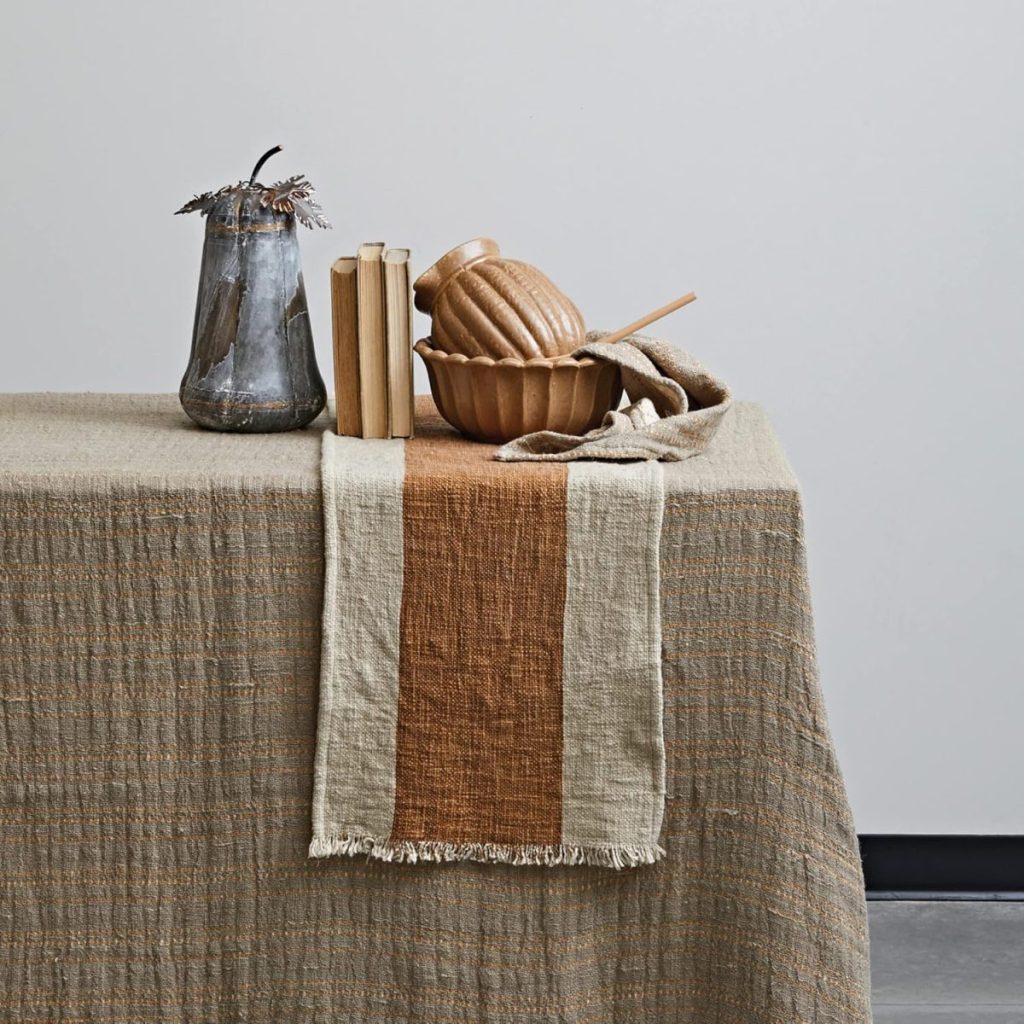 A table covered with a textured beige cloth, featuring a striped table runner, a ceramic vase, three stacked books, and a wooden bowl with a matching ladle.