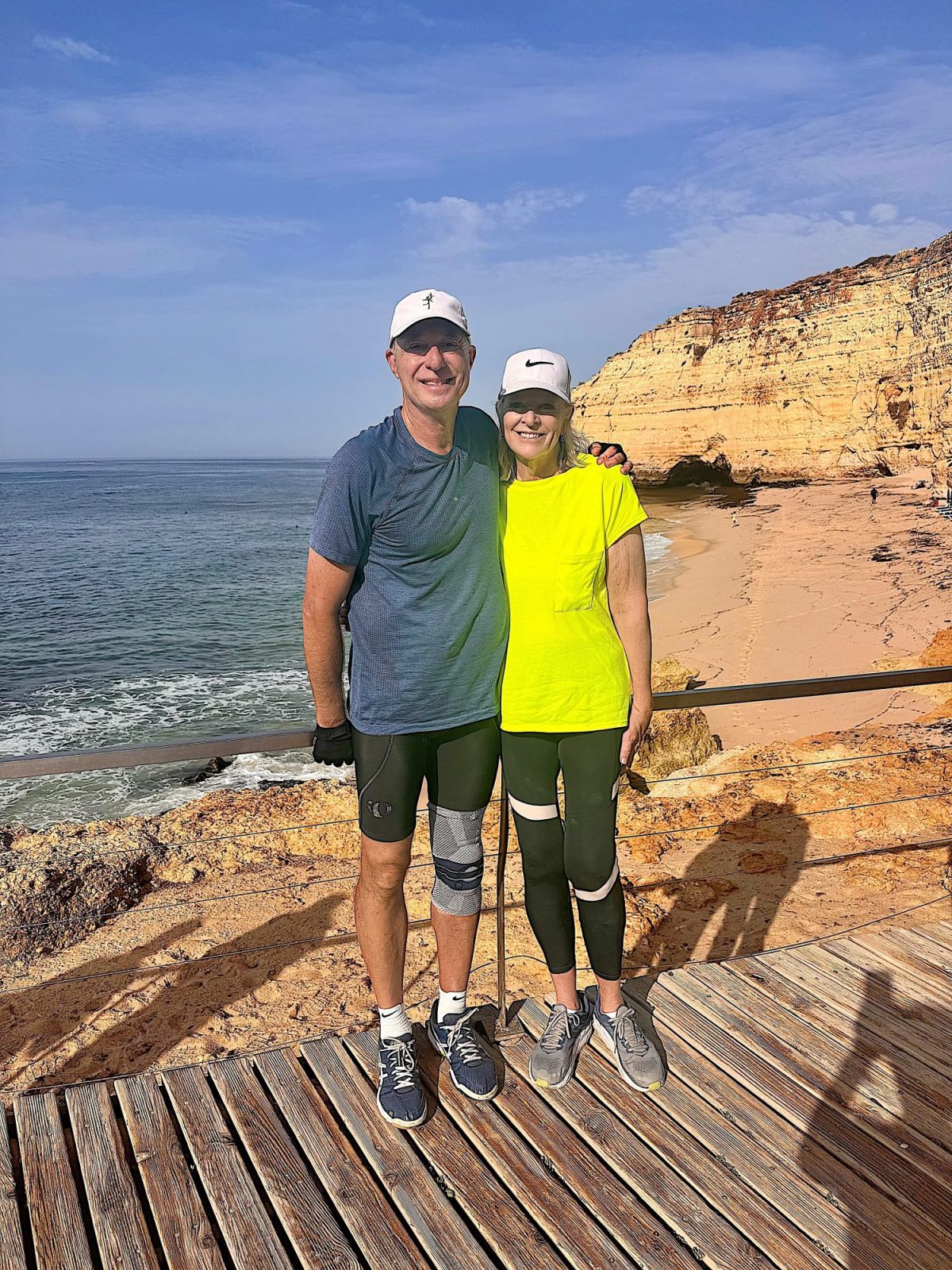 Two people in athletic wear stand on a wooden deck by the seaside with rocky cliffs and a sandy beach in the background on a sunny day.