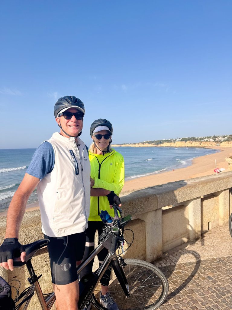 Two cyclists wearing helmets and sunglasses stand with their bikes on a seaside promenade, with a sandy beach and blue sky in the background.