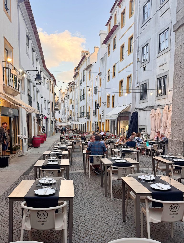 Outdoor restaurant tables set along a narrow cobblestone street with people dining, surrounded by white buildings with balconies under a partly cloudy sky.