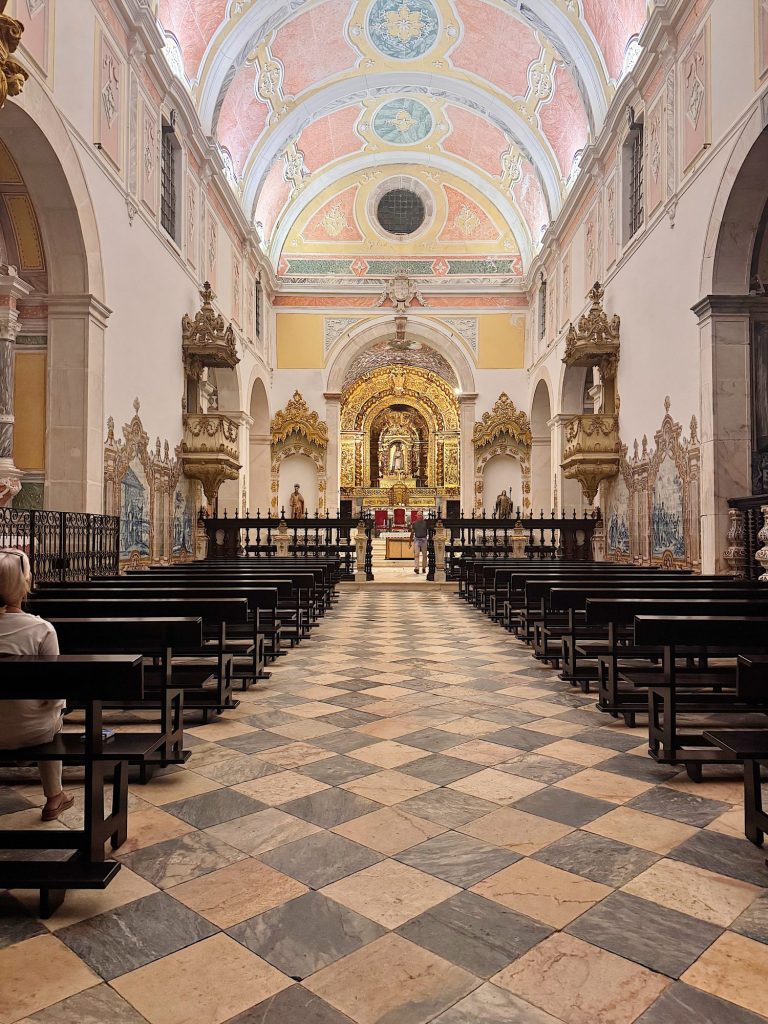 A view of a church interior with ornate altar, arched ceiling, gold decorations, tiled floor, wooden pews, and a few people inside.