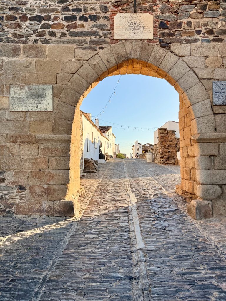 Stone archway entrance with cobblestone street leading to whitewashed buildings under a clear sky, viewed in warm sunlight.