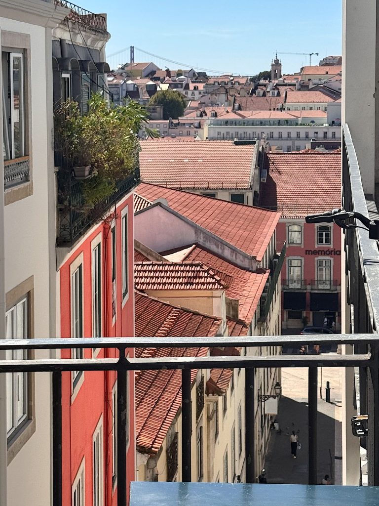 View from a balcony overlooking a narrow street lined with red-roofed buildings, with a distant bridge and cityscape visible under a clear sky.