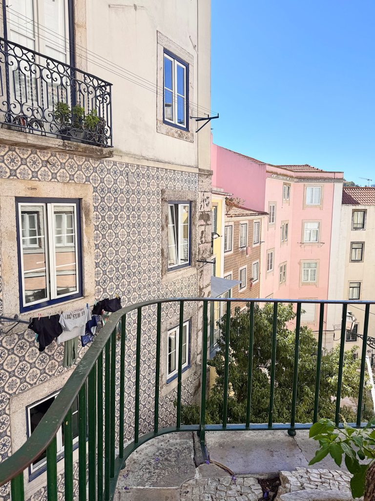 View from a balcony overlooking patterned tiled buildings, windows, and a tree, with clothes drying on a line and a clear blue sky.