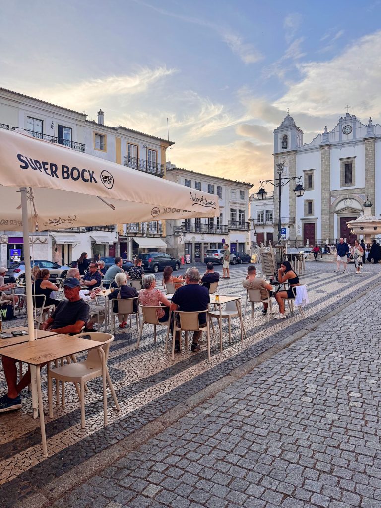 People sit at outdoor tables of a café on a cobblestone street in front of a white church with a bell tower during sunset.