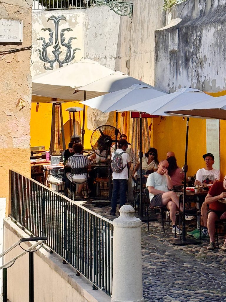 Outdoor café with several people sitting at tables under umbrellas along a yellow wall, on a cobblestone street with a railing in the foreground.