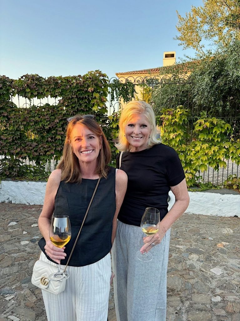 Two women stand outdoors holding glasses of white wine, smiling at the camera. Ivy-covered walls and greenery are visible in the background.