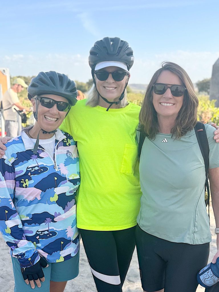 Three women wearing cycling helmets and sunglasses stand outdoors, smiling at the camera. One wears a bright yellow top, while the others wear a patterned jacket and a light green shirt.