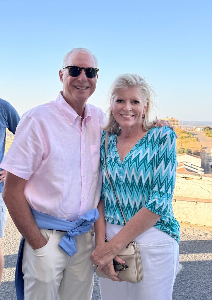 A man and a woman stand side by side outdoors on a sunny day, smiling at the camera. The man wears sunglasses and a pink shirt; the woman wears a teal patterned blouse.