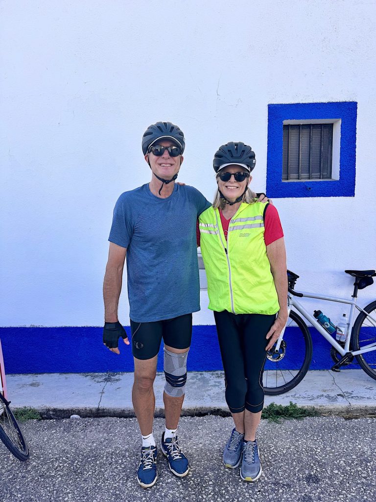 Two cyclists wearing helmets and sunglasses stand side by side in front of a white wall with blue trim. Both are smiling, and bicycles are visible in the background.