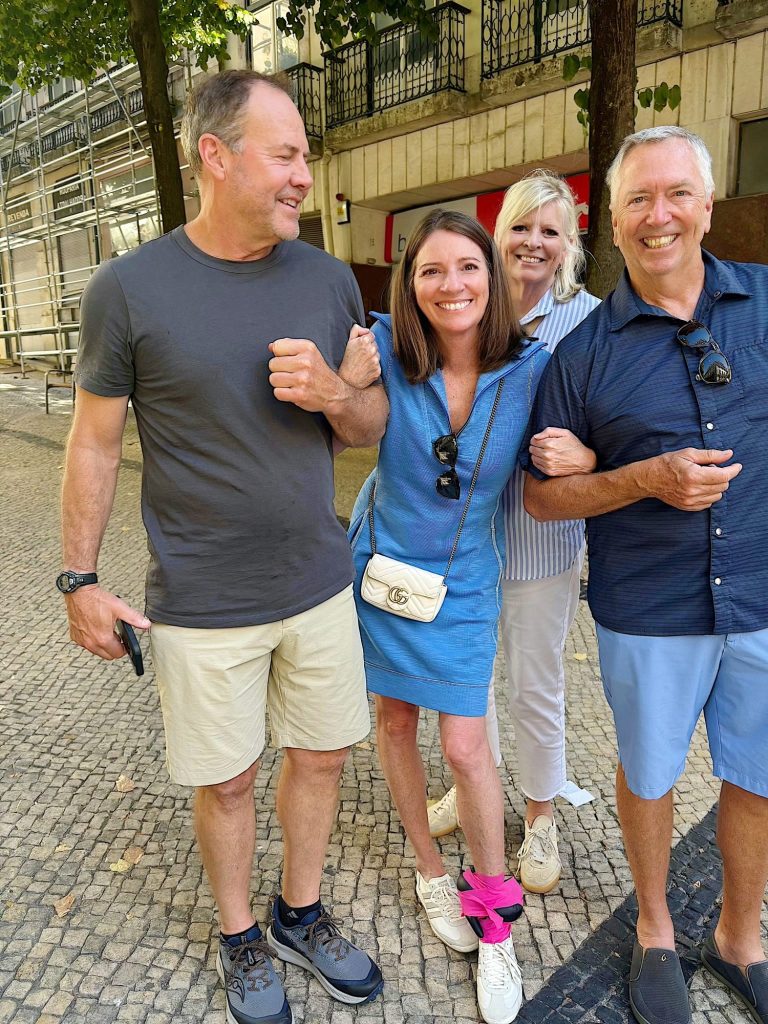 Four adults stand outdoors on a tiled walkway, smiling at the camera and linking arms. One woman has a pink cloth tied around her ankle. Trees and buildings are visible in the background.