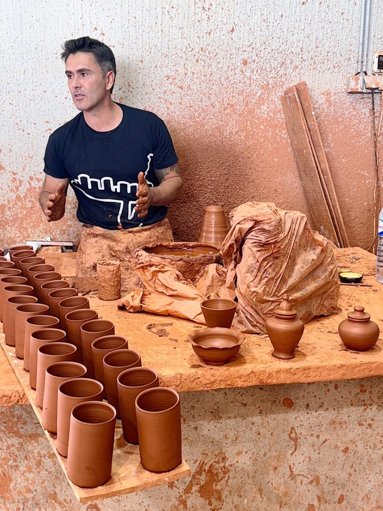 A man stands at a worktable with rows of clay cups and pots, explaining pottery-making; a large chunk of clay and pottery tools are also on the table.