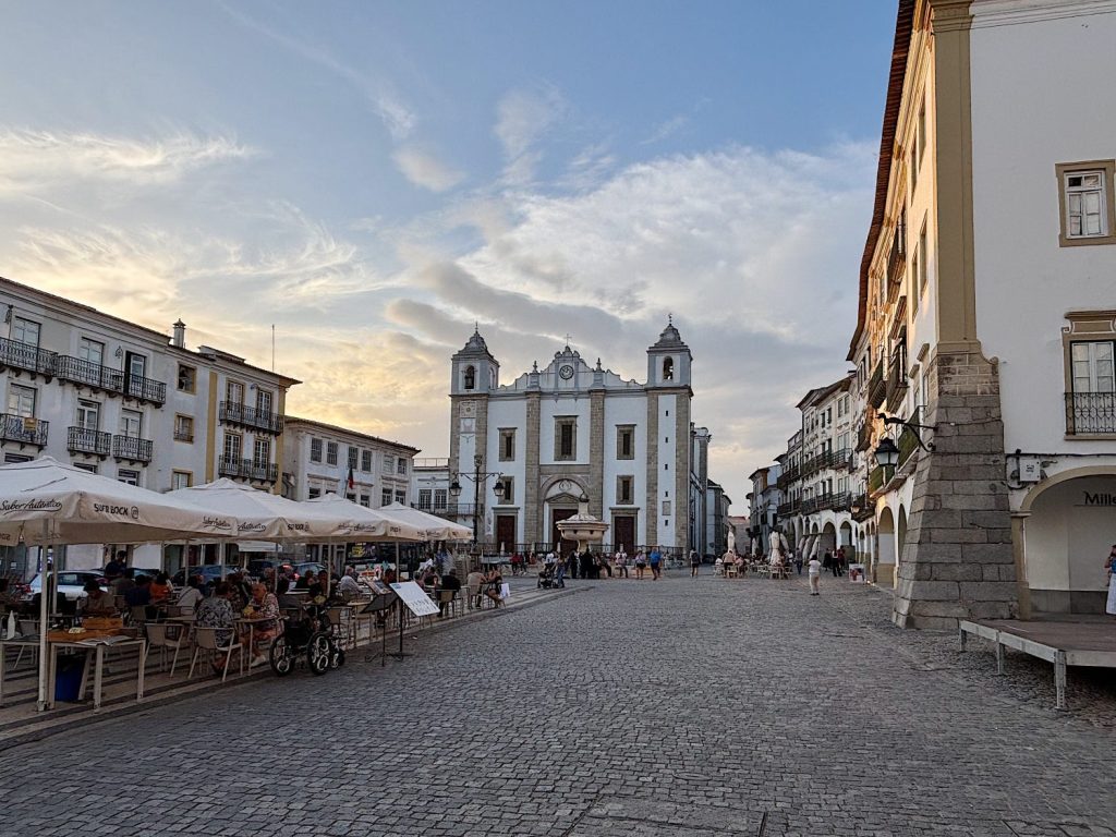 A cobblestone square with outdoor café seating and people, bordered by historic white buildings and a large church with two towers in the background under a partly cloudy sky.