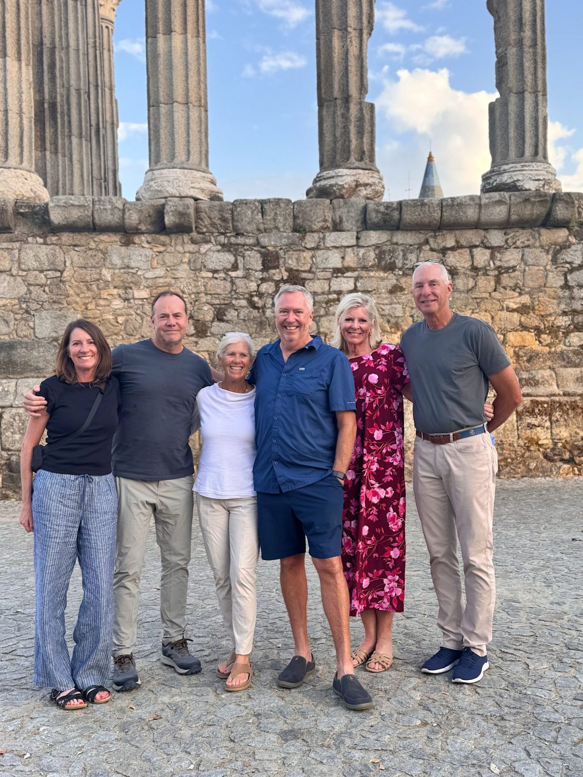 Six adults stand together smiling in front of ancient stone columns and a stone wall, posing outdoors on a sunny day.