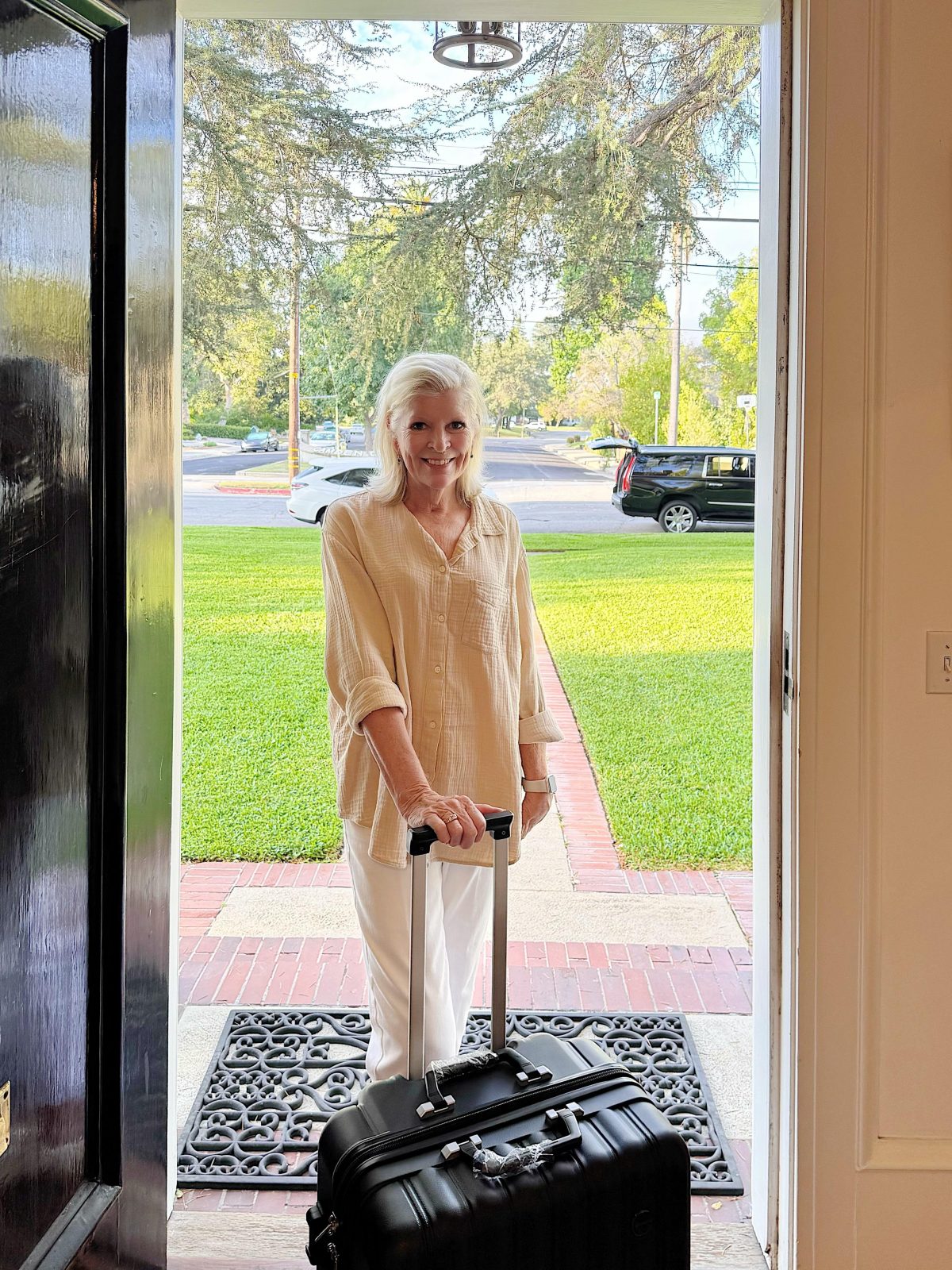 A woman stands in a doorway with a suitcase, gazing outside toward a green lawn and parked cars, ready to begin her journey to Portugal.
