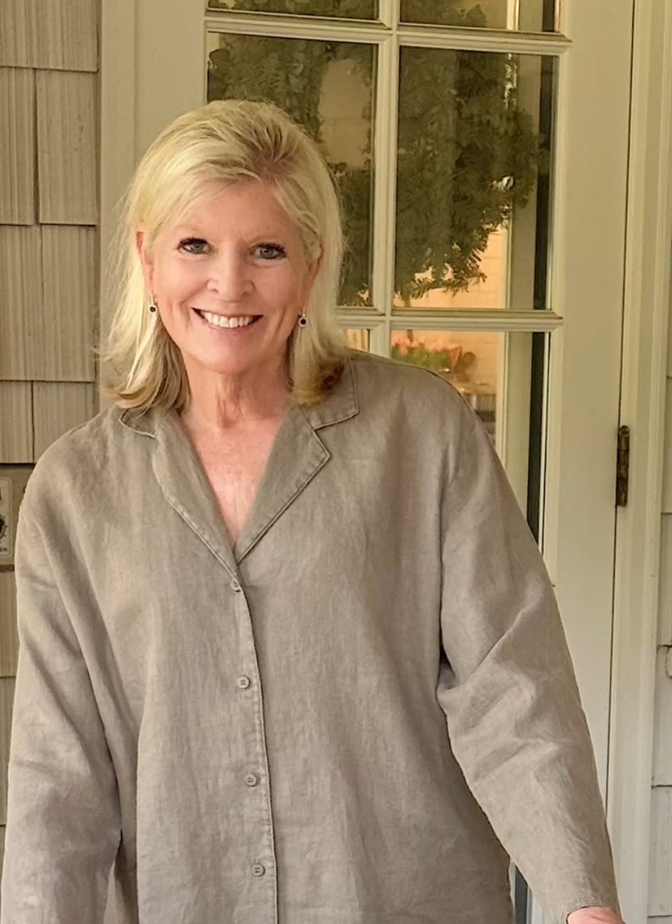 A woman with blonde hair wearing a beige button-up shirt stands and smiles in front of a glass-paned door, capturing the warm, relaxed spirit of Portugal.