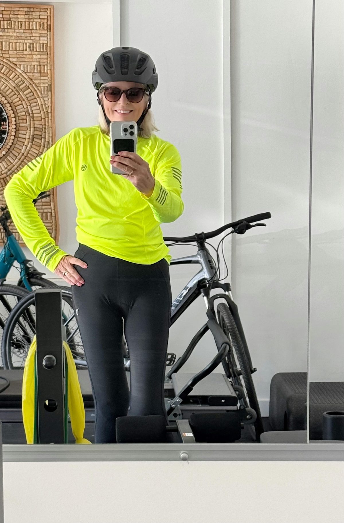 Person wearing a yellow cycling top, black helmet, and sunglasses stands indoors in Portugal, taking a mirror selfie with a bicycle and exercise equipment visible in the background.