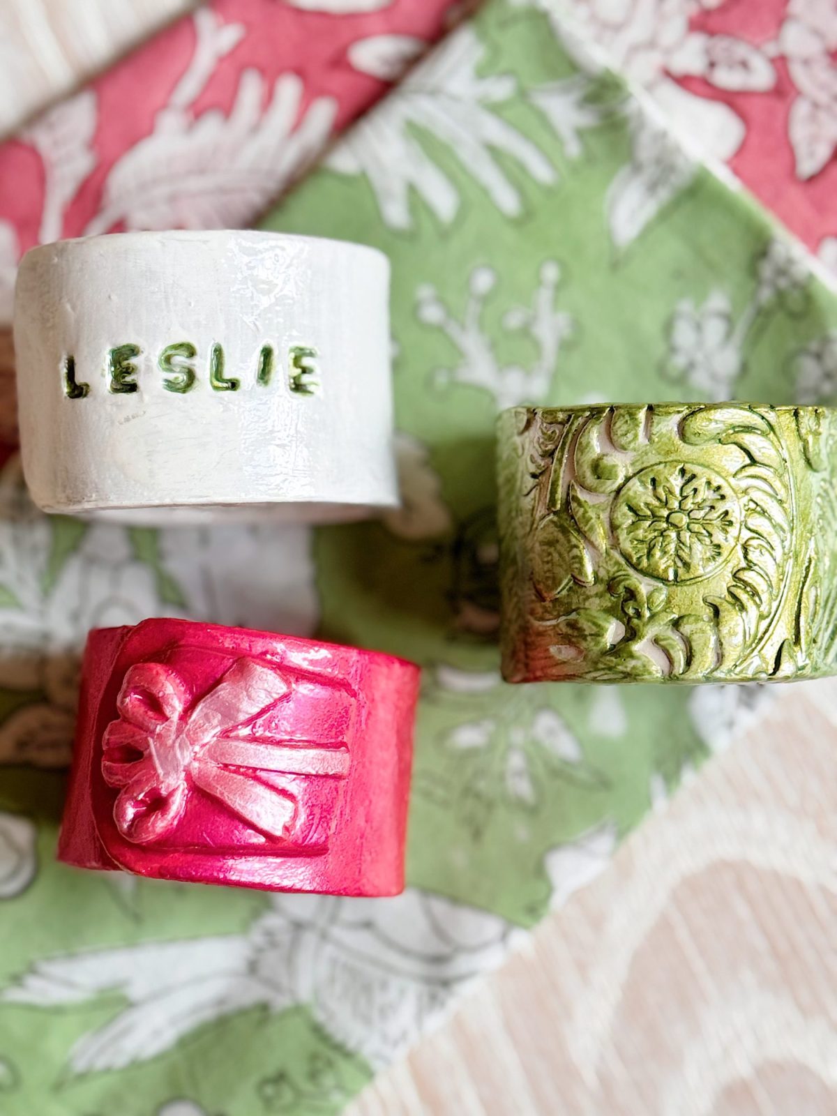 Three decorative ceramic napkin rings: one white with "LESLIE" in green letters, one green with a floral pattern, and one red with a bow design, on patterned napkins.