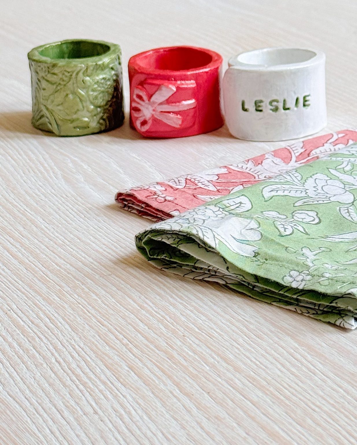 Three ceramic napkin rings&mdash;green, red, and white labeled "LESLIE"&mdash;are placed on a light wood surface next to folded patterned cloth napkins.