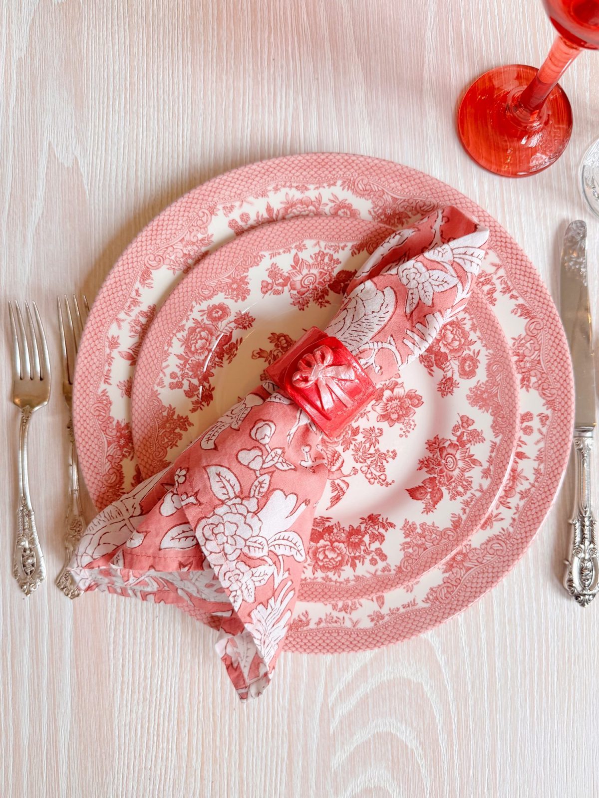 Table setting with a pink and white floral-patterned plate, matching napkin in a ring, two forks, knife, and a red glass on a light wood surface.