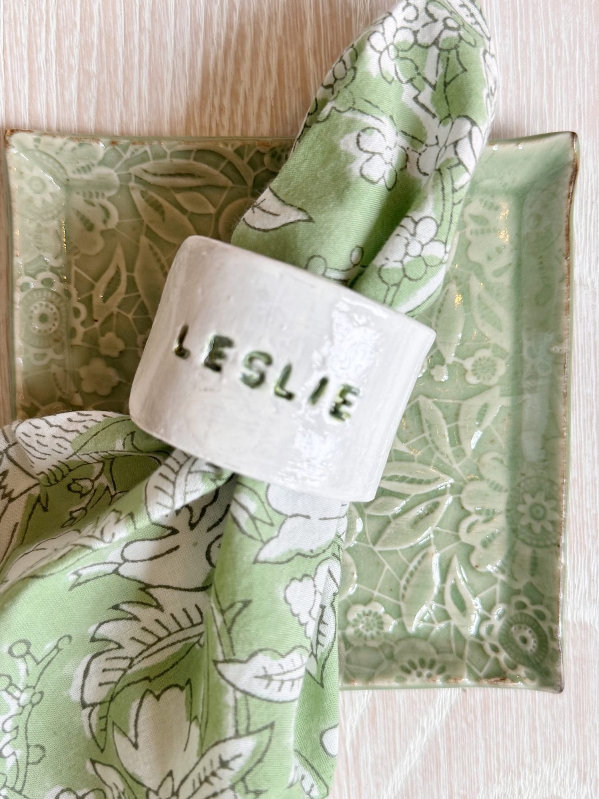 A green floral napkin with a white napkin ring labeled "LESLIE" rests on a light green patterned square plate.
