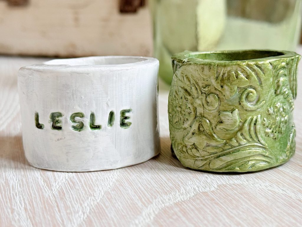 Two handcrafted napkin rings rest on a wooden surface; one is white with the name "LESLIE" stamped on it, the other is green with an ornate, floral pattern.