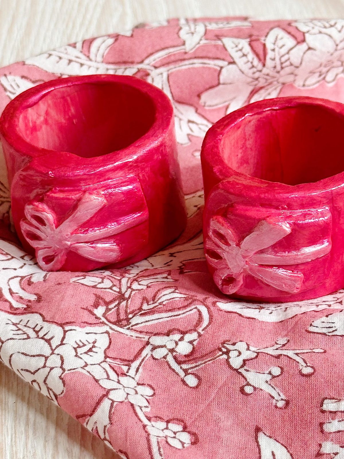 Two pink napkin rings with white bow designs are placed on a floral-patterned pink and white napkin on a light wood surface.