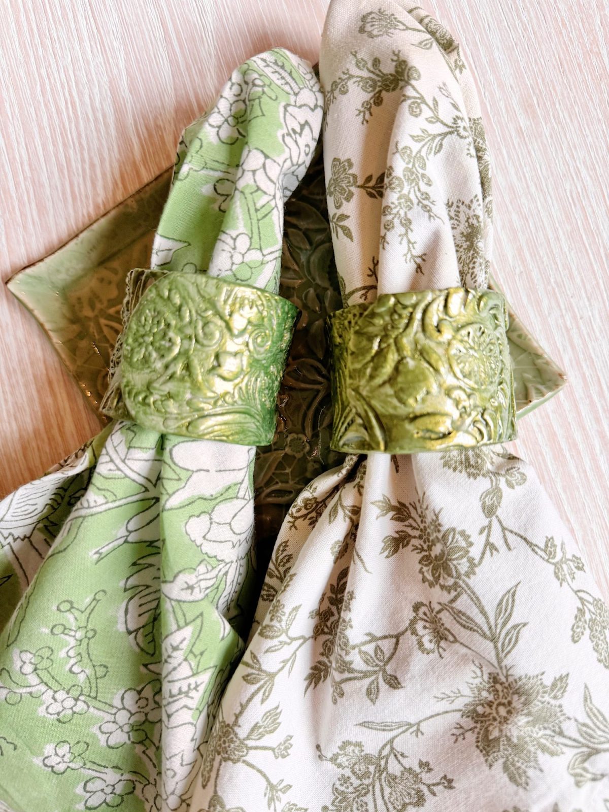Two floral-patterned cloth napkins, one green and one white, are held by ornate gold napkin rings on a light-colored wooden surface.