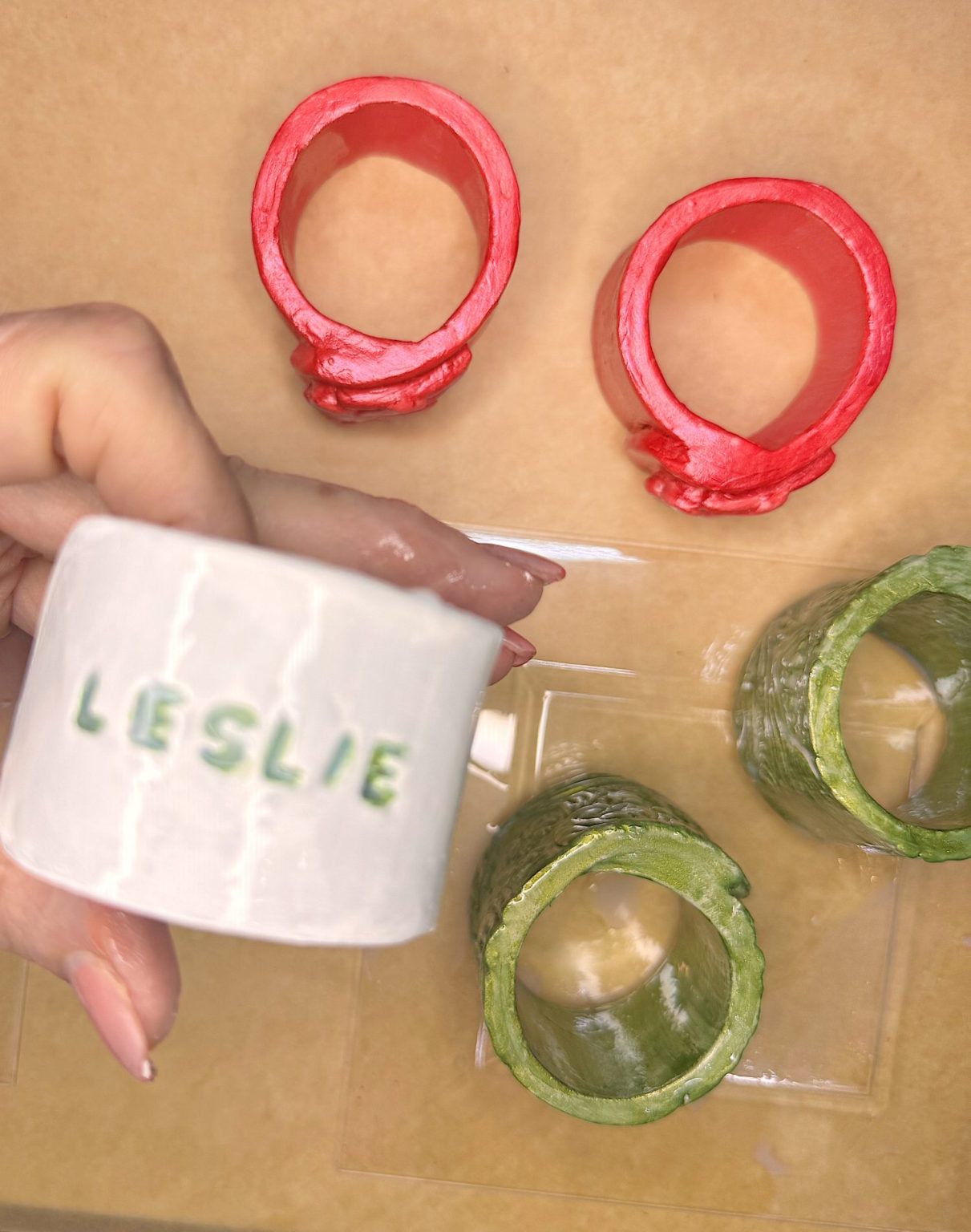 A hand holds a white ceramic napkin ring labeled "LESLIE"; two red and two green napkin rings are placed on a beige surface below.