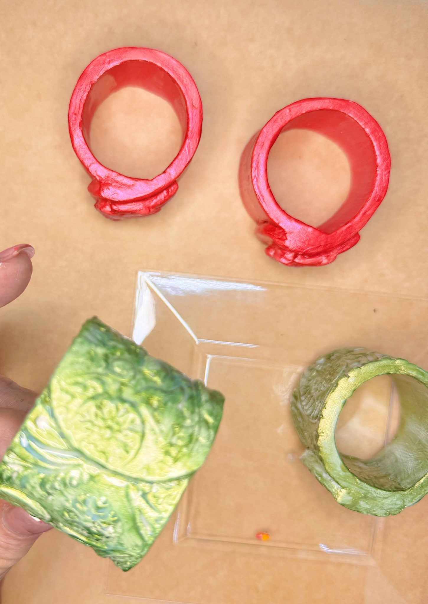 Three ornate bangle molds, two red and one green, are displayed on a beige surface with a clear square tray. A hand holds the green bangle mold.