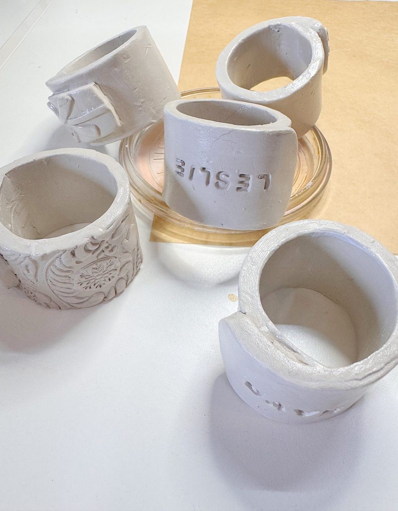 Five unfinished, white clay napkin rings with different textures and designs are arranged on a white surface near a glass dish and a piece of brown paper.
