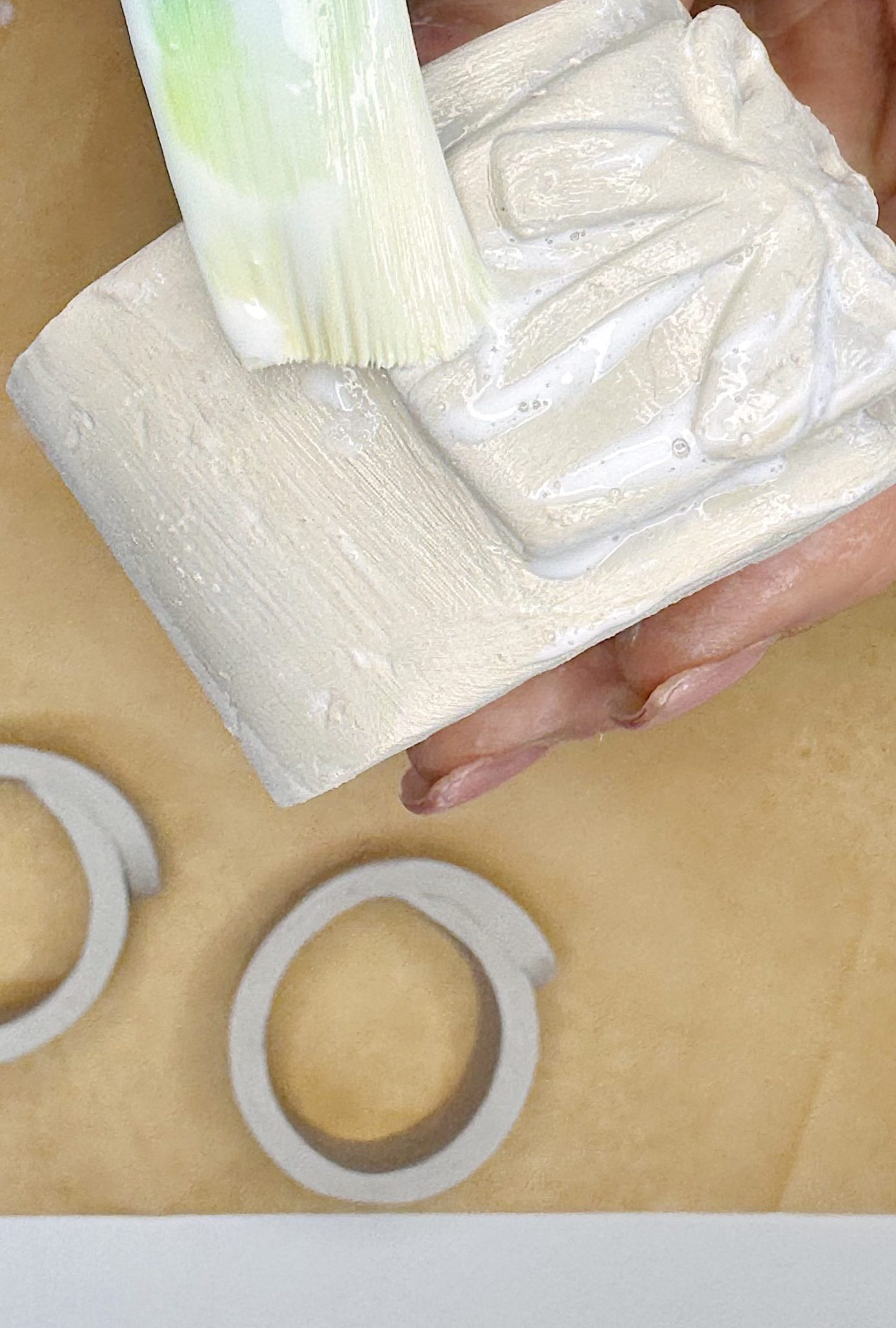 A hand applies white paint with a brush to a textured, rectangular object while two gray circular items lie on a brown surface nearby.