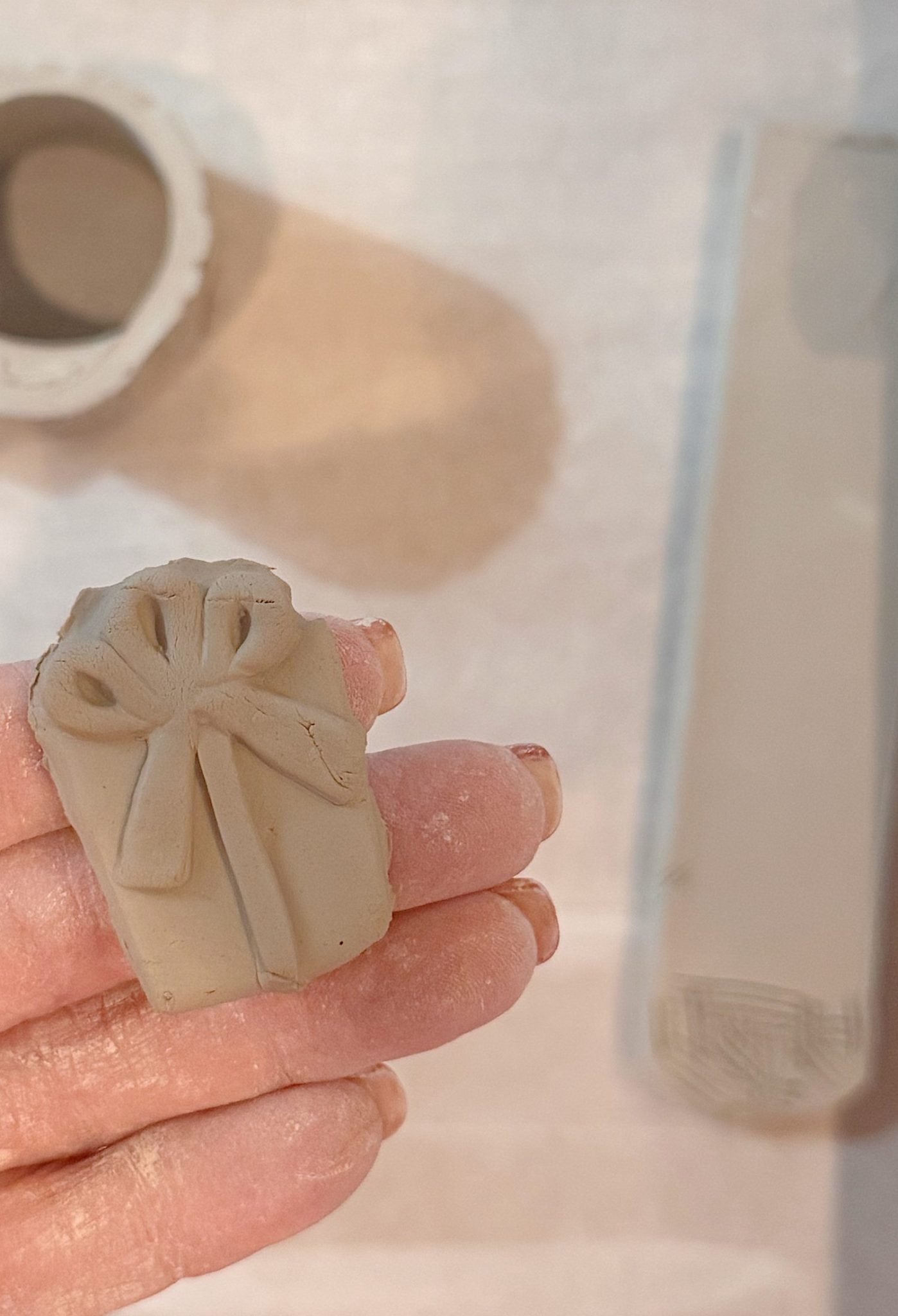 A hand holds a small piece of clay shaped like a gift with a bow, with clay tools and a container visible in the background.