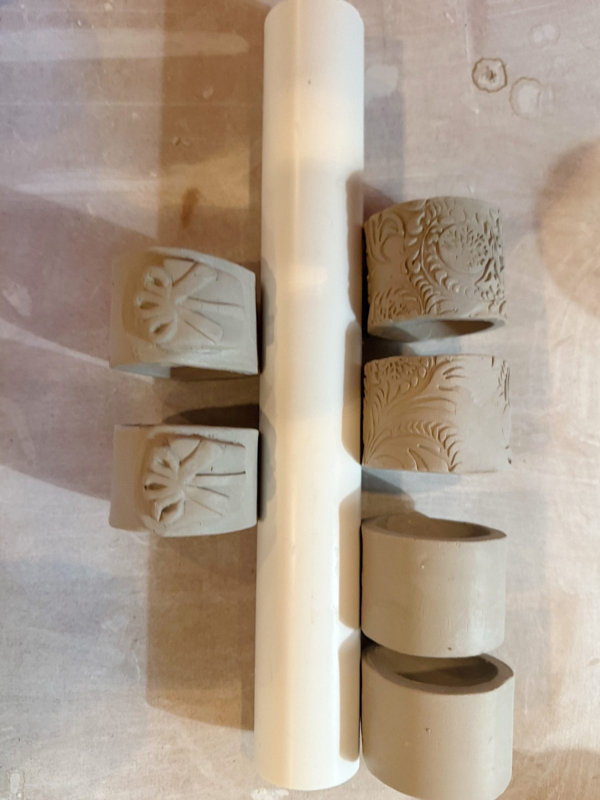 Six small clay cylinders with various patterns are arranged around a white rolling pin on a light-colored surface.