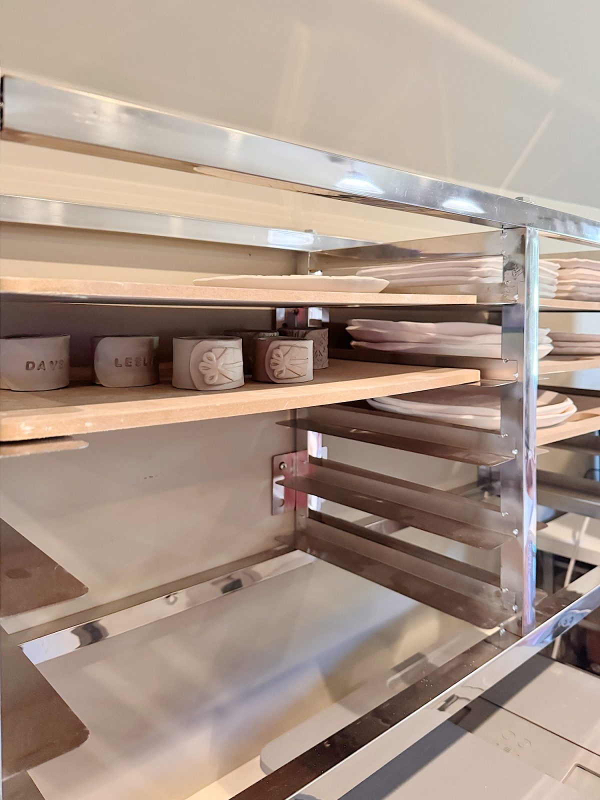 Metal drying rack holding several shelves with unfinished ceramic pieces, including small cylindrical objects and flat plates, in a pottery studio setting.