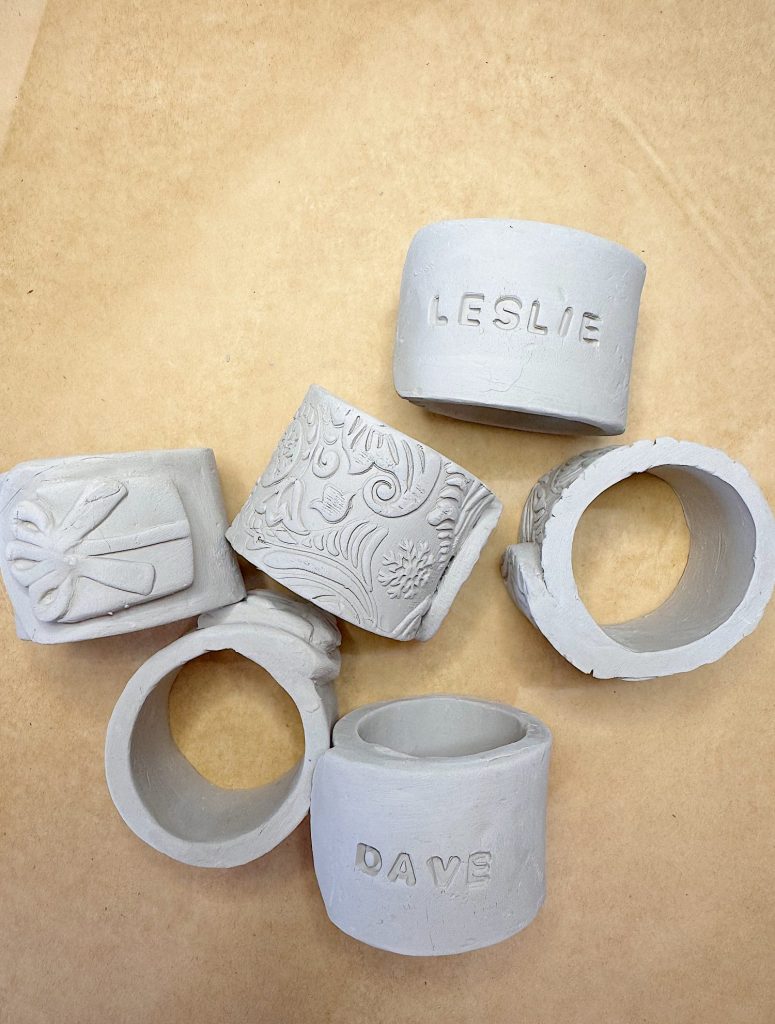 Six unglazed ceramic napkin rings, some engraved with names ("LESLIE" and "DAVE") and textured patterns, are arranged on a tan surface.
