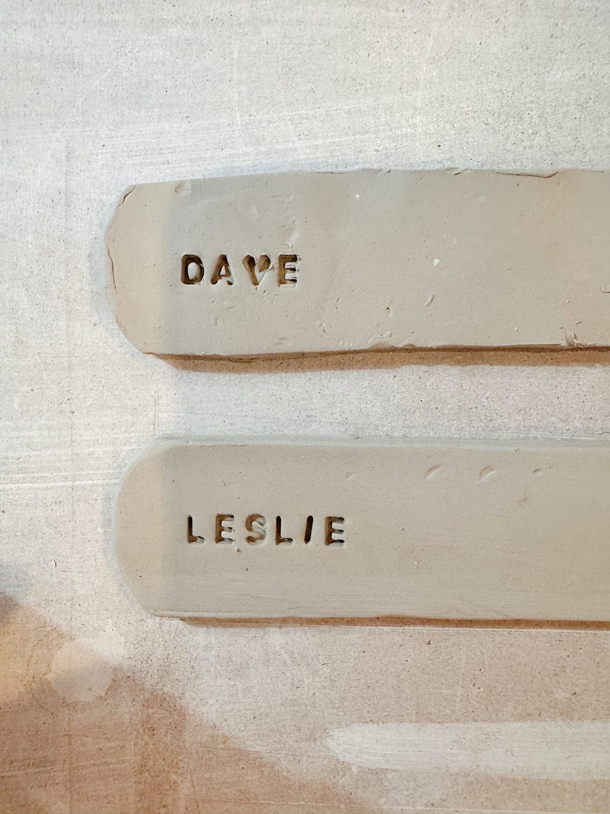 Two clay strips with the names "DAVE" and "LESLIE" stamped into them, placed on a light-colored surface.