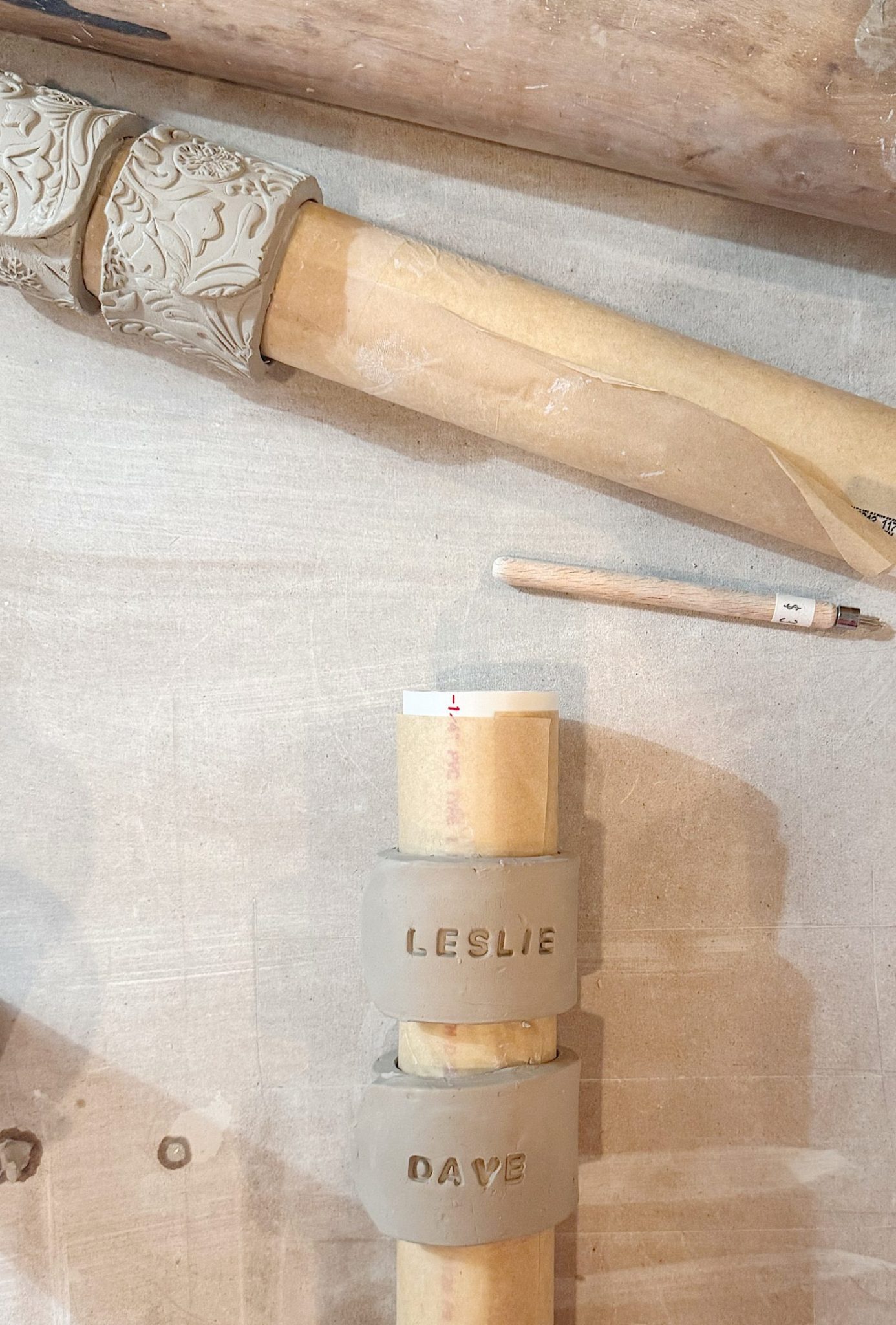 Two clay rollers are wrapped in brown paper on a work surface; one has a floral pattern, the other has clay rings labeled "LESLIE" and "DAVE." A sculpting tool is also visible.