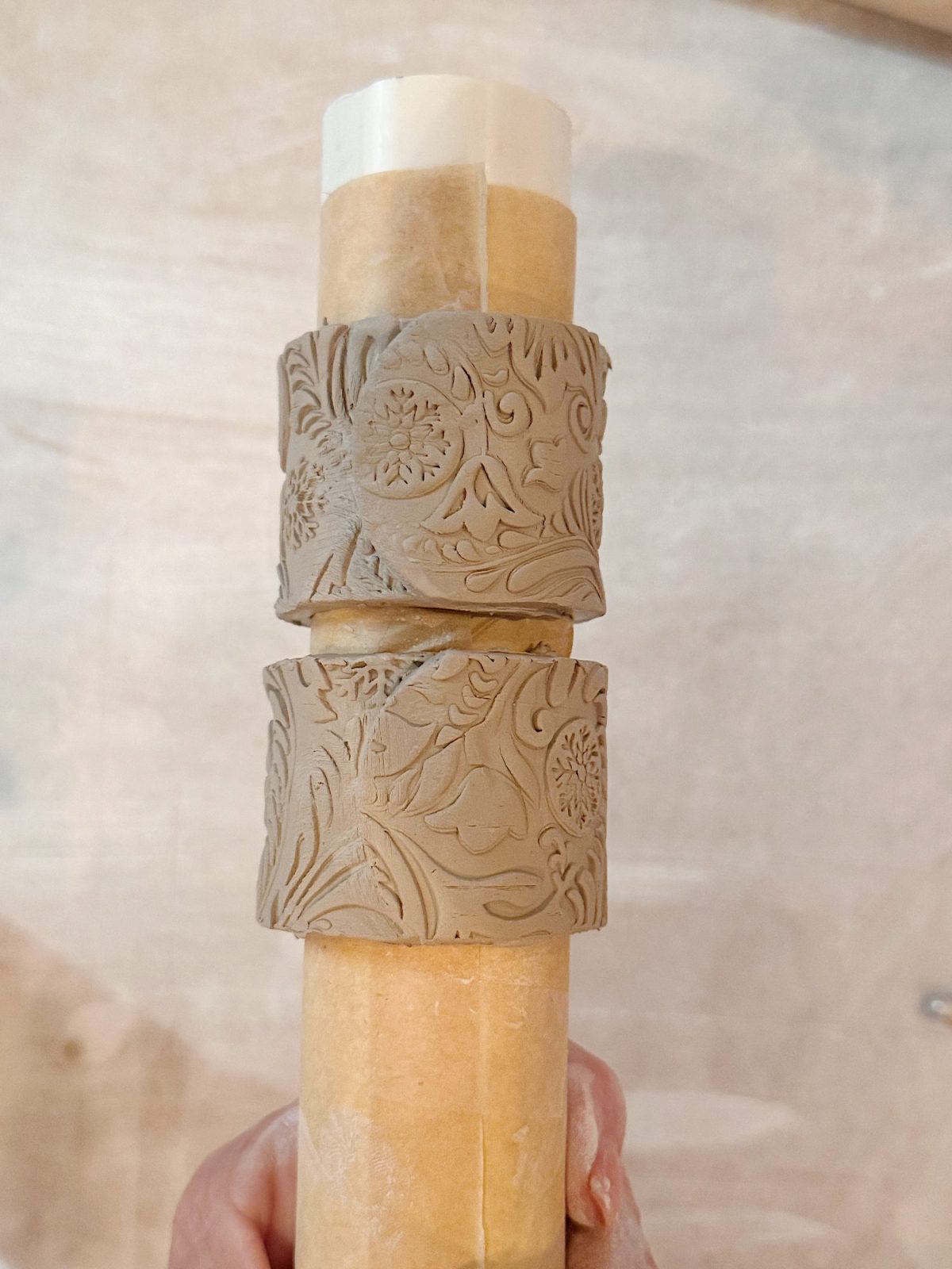 A hand holds a cylindrical rolling pin with two textured clay rings featuring intricate floral and leaf patterns placed around it.