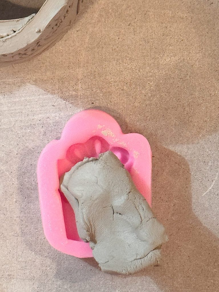 A pink plastic cookie cutter shaped like a paw print is partially filled with gray modeling clay on a cardboard surface.