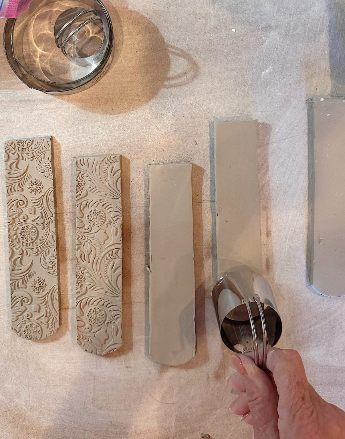 A hand uses a round metal cutter on a slab of clay beside five clay strips, two with floral patterns and three with smooth surfaces, on a work surface.