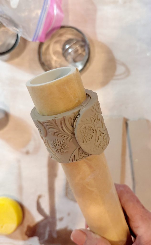 A hand holds a rolling pin wrapped with a piece of clay that has an ornate, floral pattern pressed into its surface.