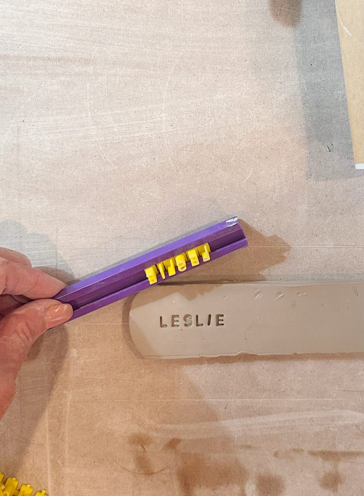A hand holds a purple stamp tool with yellow letters over a clay slab with the word "LESLIE" stamped into it.