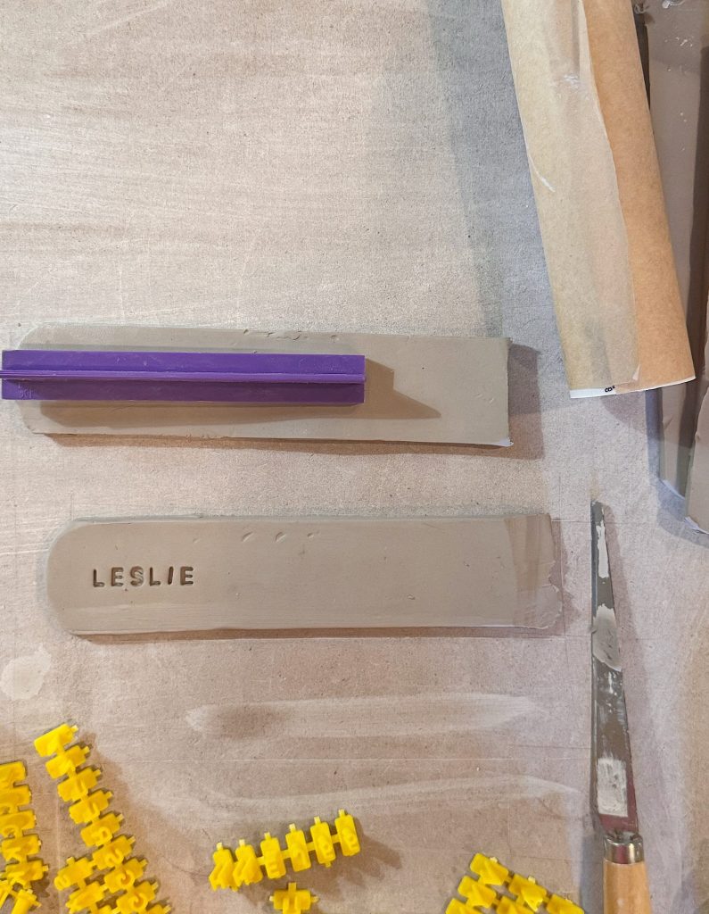 Two rectangular clay slabs on a table, one stamped with the name "LESLIE" and the other with a purple tool resting on top. Yellow plastic pieces and a metal tool are nearby.