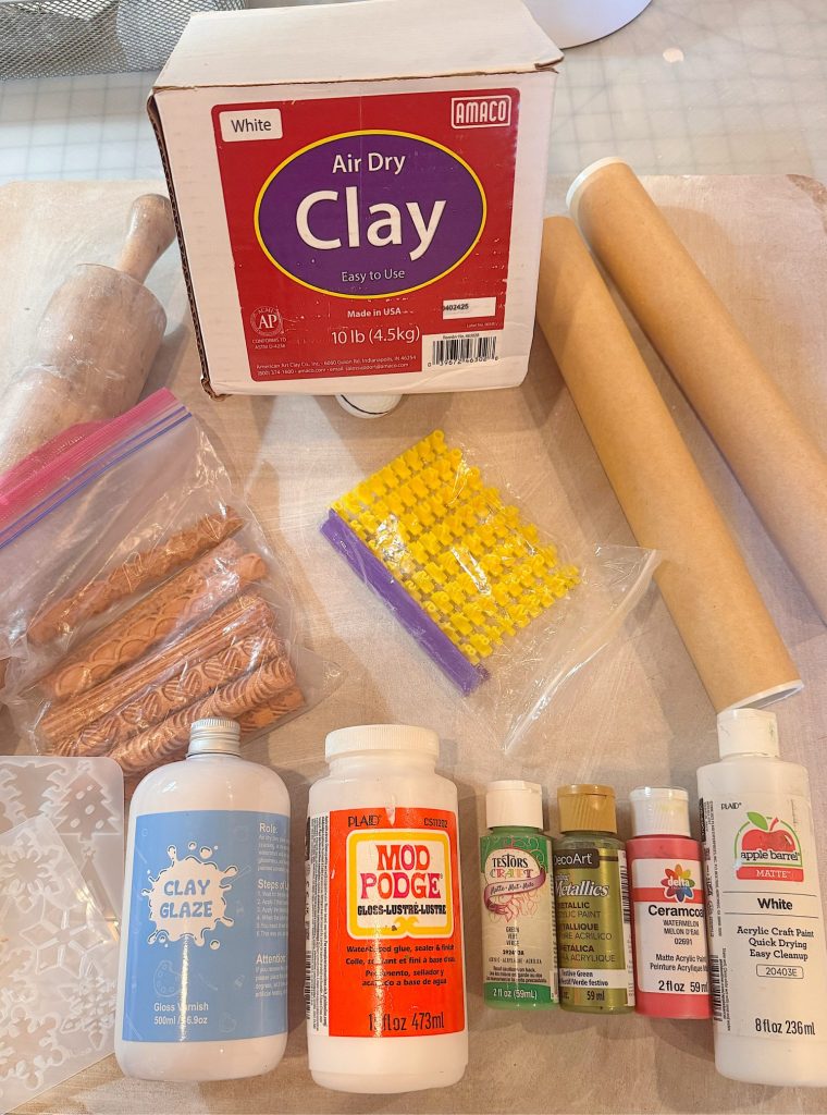 Art and craft supplies laid out, including a box of air dry clay, rolling pins, bottles of glaze and paint, Mod Podge, stamps, texture sheets, and small yellow beads.